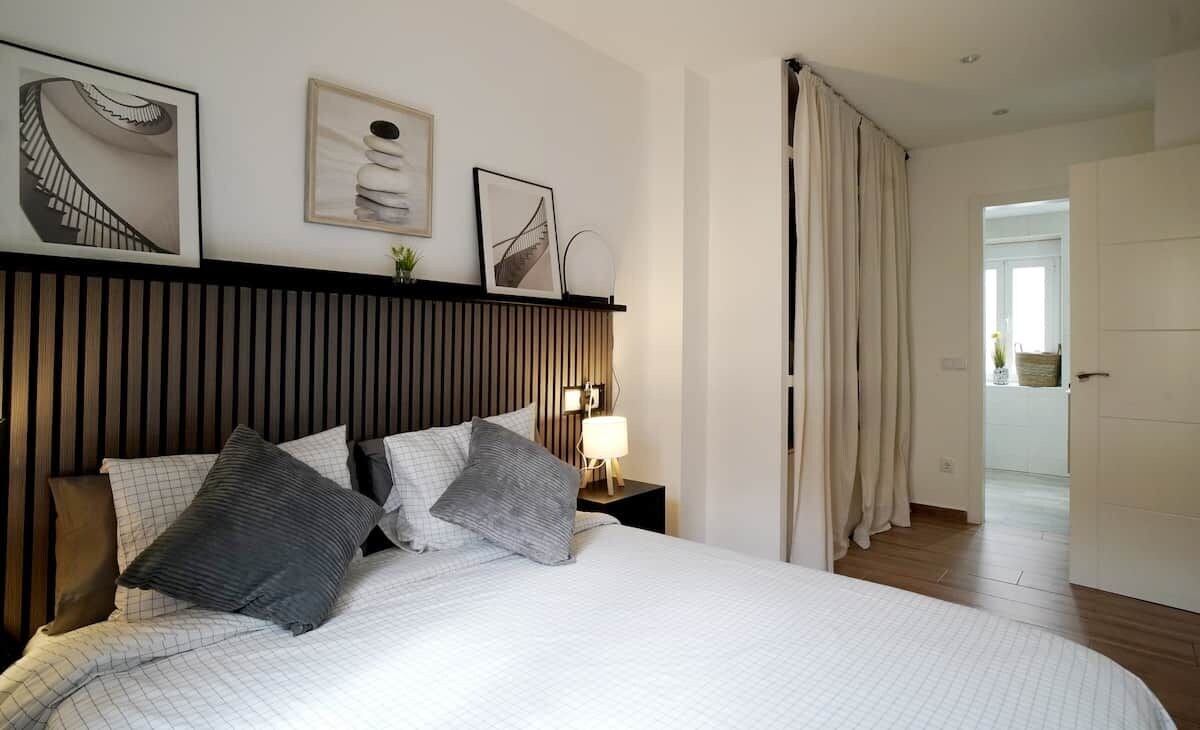 Habitación de hotel con cama, mesita de noche con lámpara, estante en la pared con fotografías y vista al pasillo.
