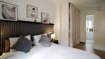 Habitación de hotel con cama, mesita de noche con lámpara, estante en la pared con fotografías y vista al pasillo.