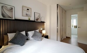 A hotel room with a bed, a nightstand with a lamp, a wall-mounted shelf with pictures, and a view of the hallway. - Murallas Home (Ávila)