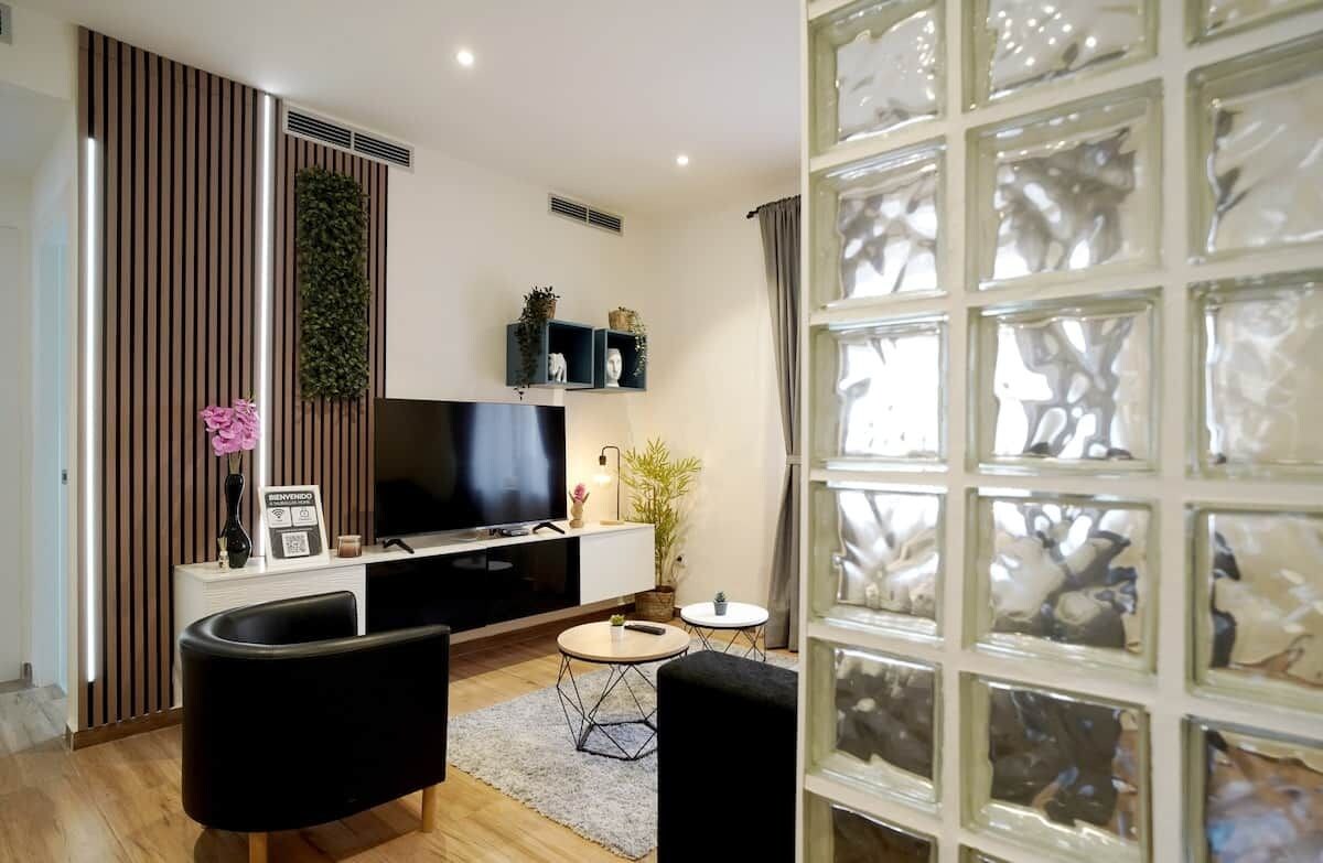 A modern living room with a flat-screen TV, black sofas, a coffee table, and a decorative plant.