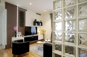 A modern living room with a flat-screen TV, black sofas, a coffee table, and a decorative plant.