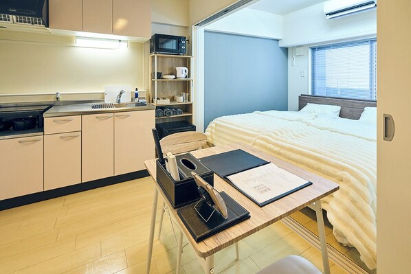 A compact hotel room with a kitchenette, a bed with white bedding, a desk with a laptop and documents, and a window with blinds.