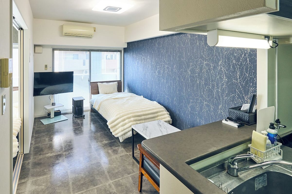 Habitación de hotel con cama, escritorio con lavamanos, televisor y ventana con persianas.