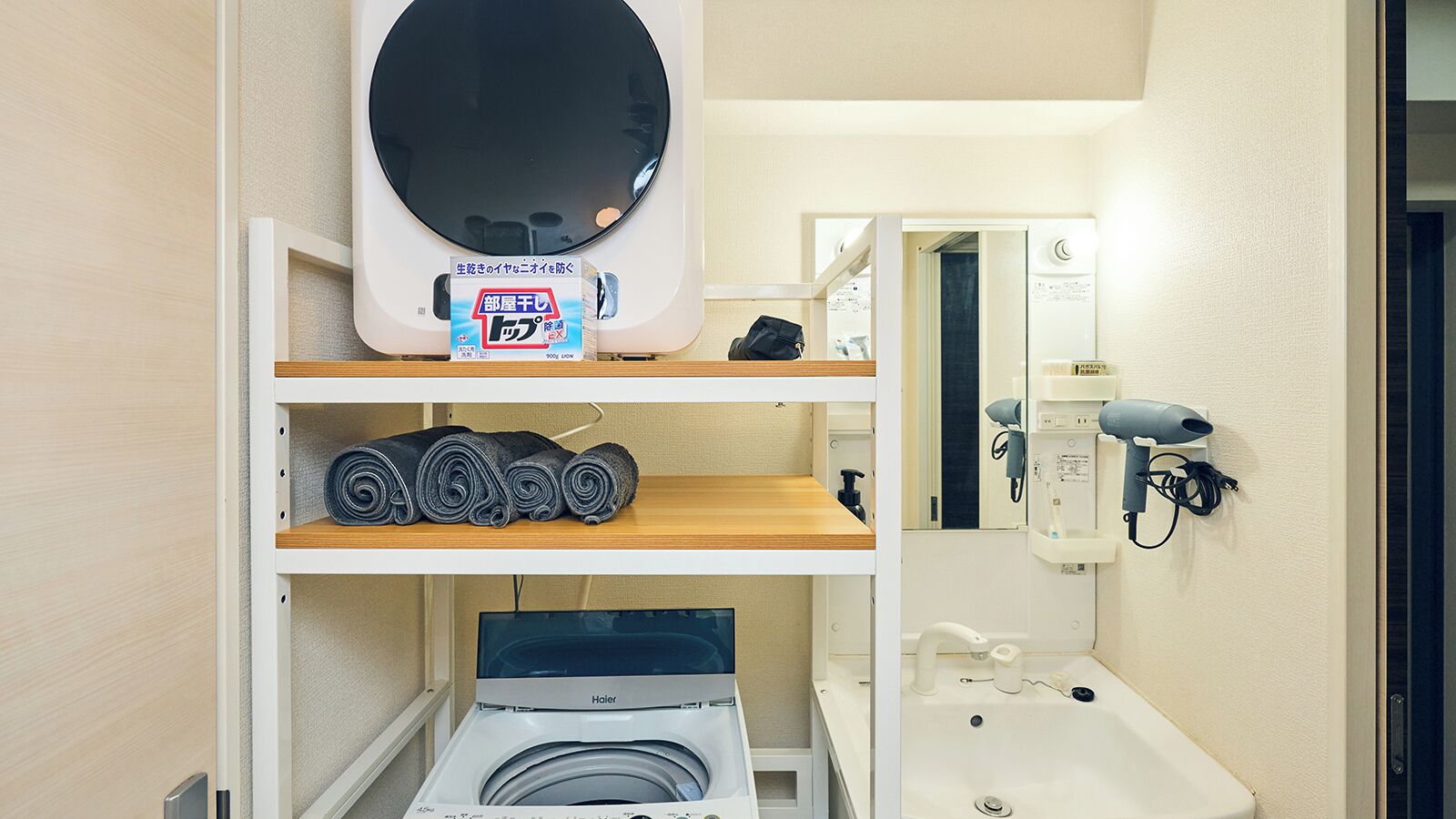 A compact hotel bathroom with a washing machine, a hairdryer, and neatly rolled towels on shelves.