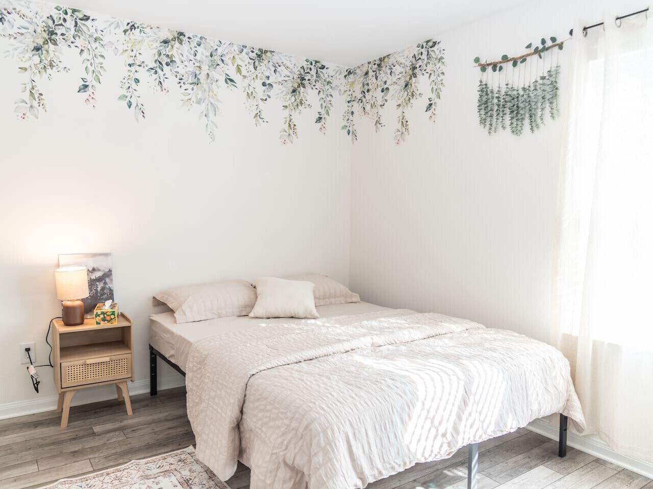 Een slaapkamer met een bed, nachtkastje, lamp en een plant aan de muur.
