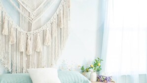 Cama com um painel de macramé na parede, mesa de cabeceira com uma planta e janela com cortinas finas.