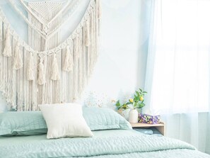 Ein Bett mit Wandbehängung aus Makramee, ein Nachttisch mit Pflanze und ein Fenster mit durchscheinenden Vorhängen.