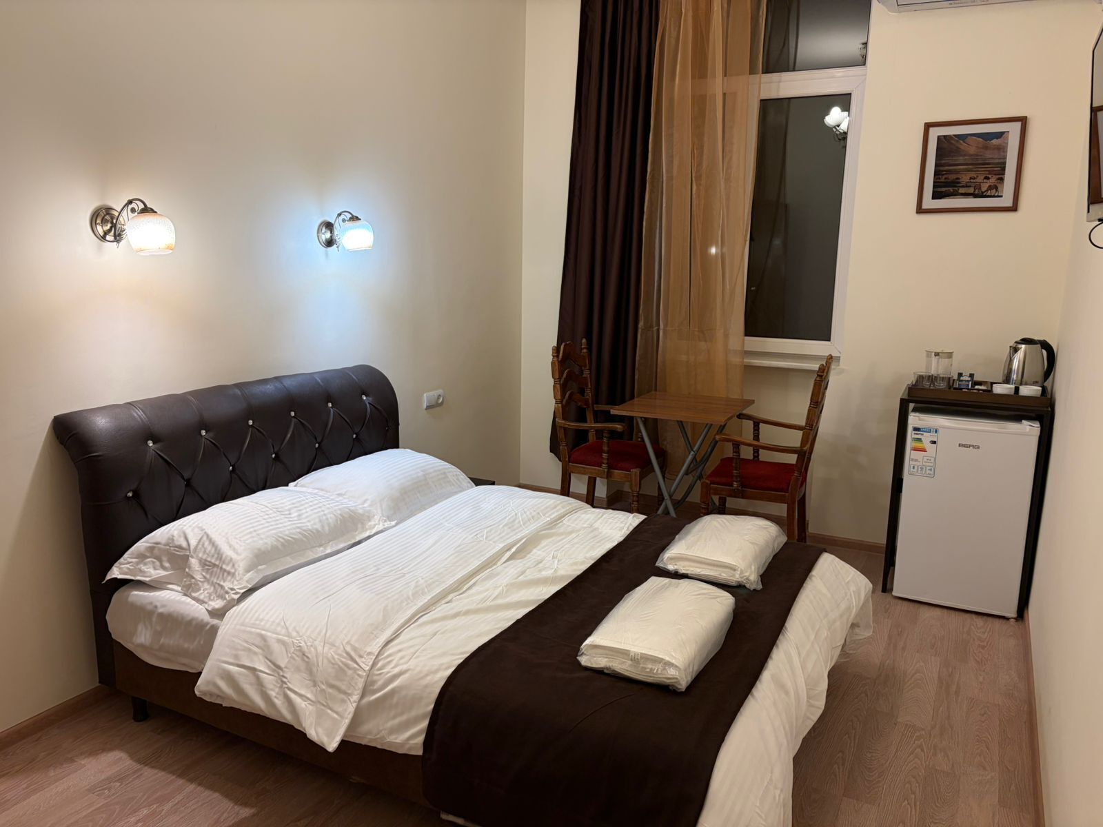 Une chambre d’hôtel avec un lit, une petite table avec des chaises, un réfrigérateur et une fenêtre avec des rideaux.
