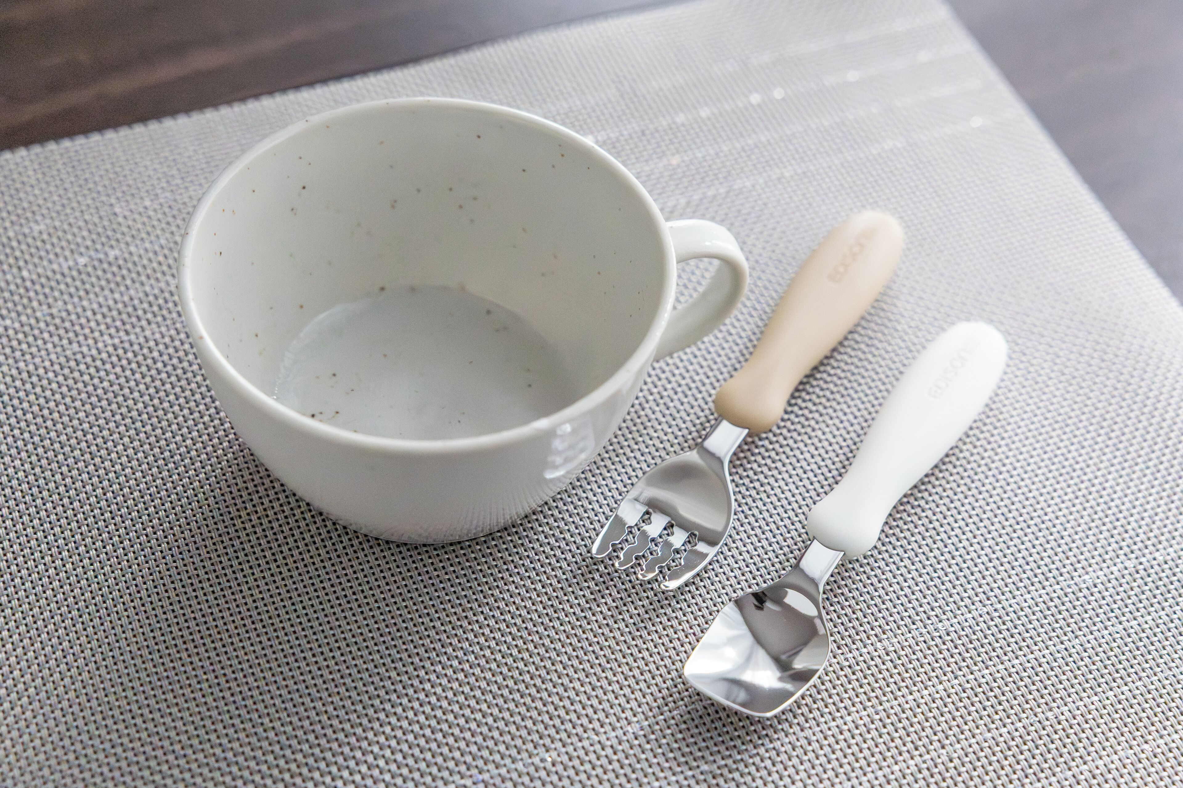A white ceramic mug with a handle and two metal utensils on a textured mat.