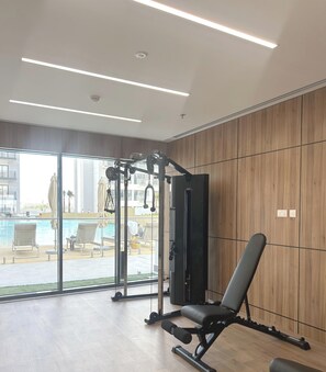 Ein Fitnessraum mit einer Bank, Kraftmaschine sowie Blick auf ein Schwimmbecken und einen Außenbereich.