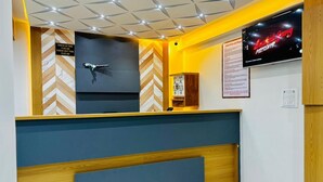 A hotel reception area with a modern design, featuring a check-in/out desk, a television displaying a movie, and a signboard with hotel information. - HOTEL HUNTER  (Bardoli)
