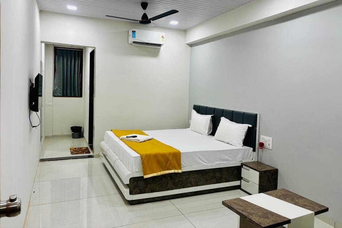 A modern hotel room with a bed, bedside tables, a ceiling fan, an air conditioning unit, and a small bathroom with a toilet and sink.