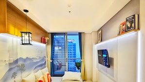A modern hotel room with a bed, a TV, a mounted TV, a wall-mounted air conditioner, and a window with a view of buildings. - Keio By SanLiving - 2BR - HubLife Mall (Jakarta)