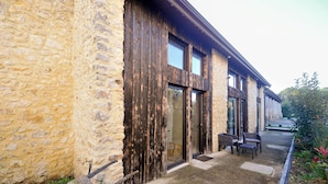 Ein Gebäude aus Stein mit großen Holztüren und -fenstern, eine gepflasterte Terrasse mit Außenbestuhlung und ein Garten.