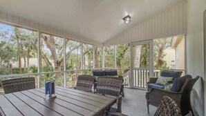 Une terrasse couverte, meublée de meubles en osier, avec une table agrémentée d’un chandelier, et de grandes fenêtres offrant une vue sur les arbres et un bâtiment.