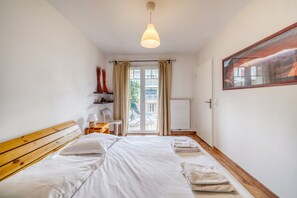 A bedroom with a wooden headboard, a bed with white linens, a bedside table with a lamp, a chair, a window with a view of the outside, and a radiator. - Esprit Bassin – Balcony, pool, and nearby beaches (Pyla sur Mer)