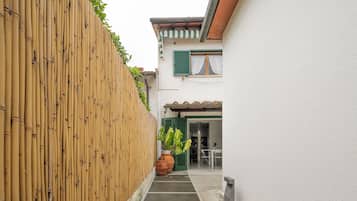 A narrow alleyway with a bamboo fence, a potted plant, and a building with green shutters and a tiled roof.