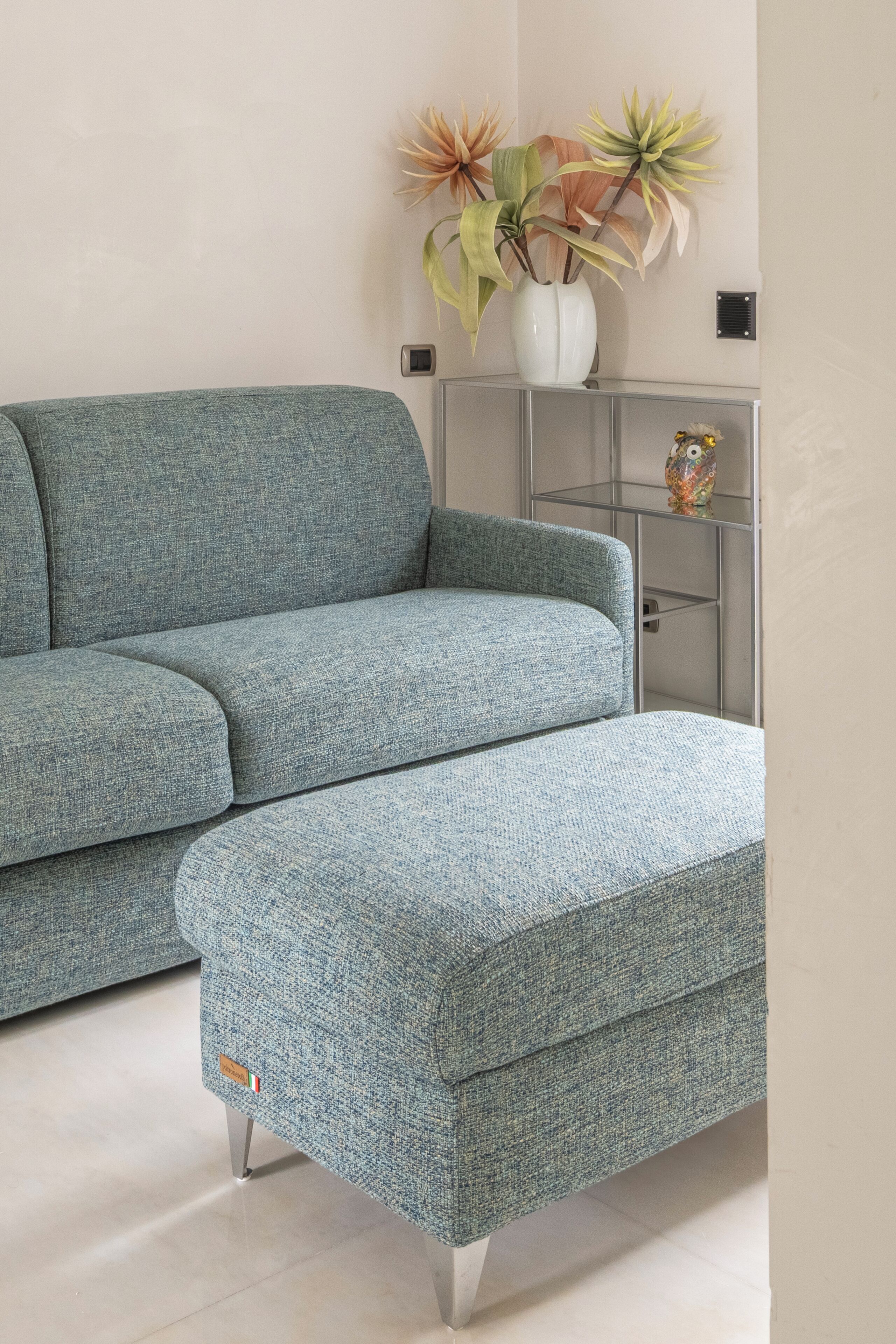 Un salon moderne avec un canapé bleu et un pouf assorti, un vase blanc contenant des fleurs séchées et une petite étagère sur laquelle est posé un objet décoratif.