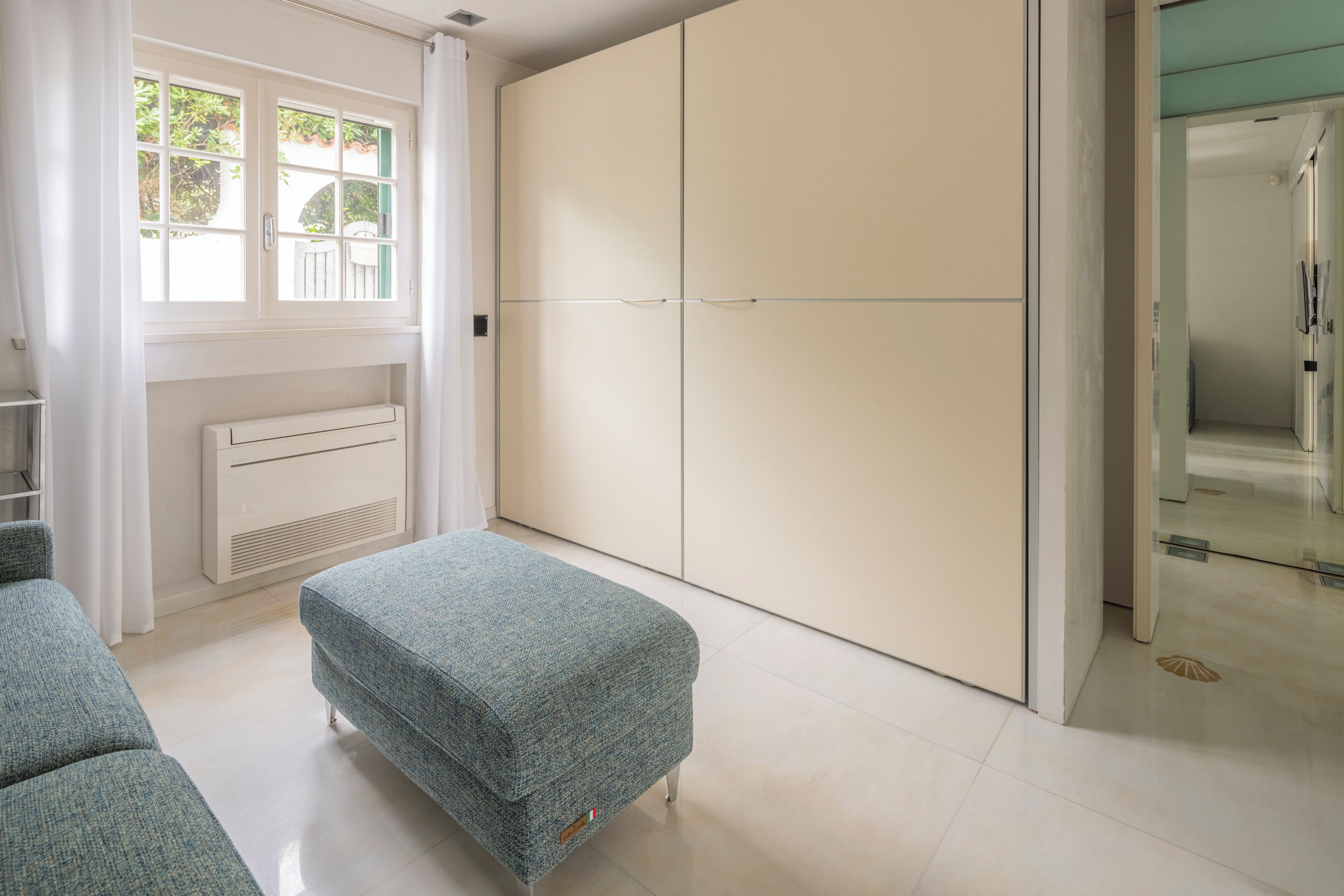 Een slaapkamer met een grote kast, een raam met gordijnen, een radiator en een klein pouf.