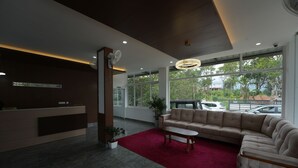 A modern reception area with a reception desk, a large beige sofa, a red carpet, and large windows offering a view of the outdoors. - Zeezmount Resorts & Spa Munnar (Devikolam)
