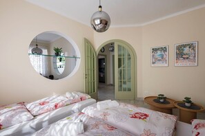 Une chambre avec deux lits, un miroir rond, un luminaire rond au plafond, une porte verte et deux horloges fixées au mur.