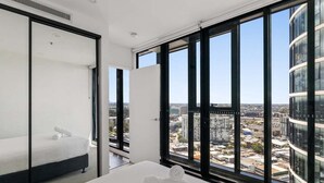 Una habitación de hotel moderna con un ventanal que ofrece vistas a la ciudad, una cama con cabecero blanco y una cama bien hecha con una toalla enrollada.
