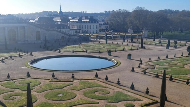 Ein formeller Garten mit einem zentralen Teich, geometrischer Bepflanzung und Gebäuden in der Ferne.