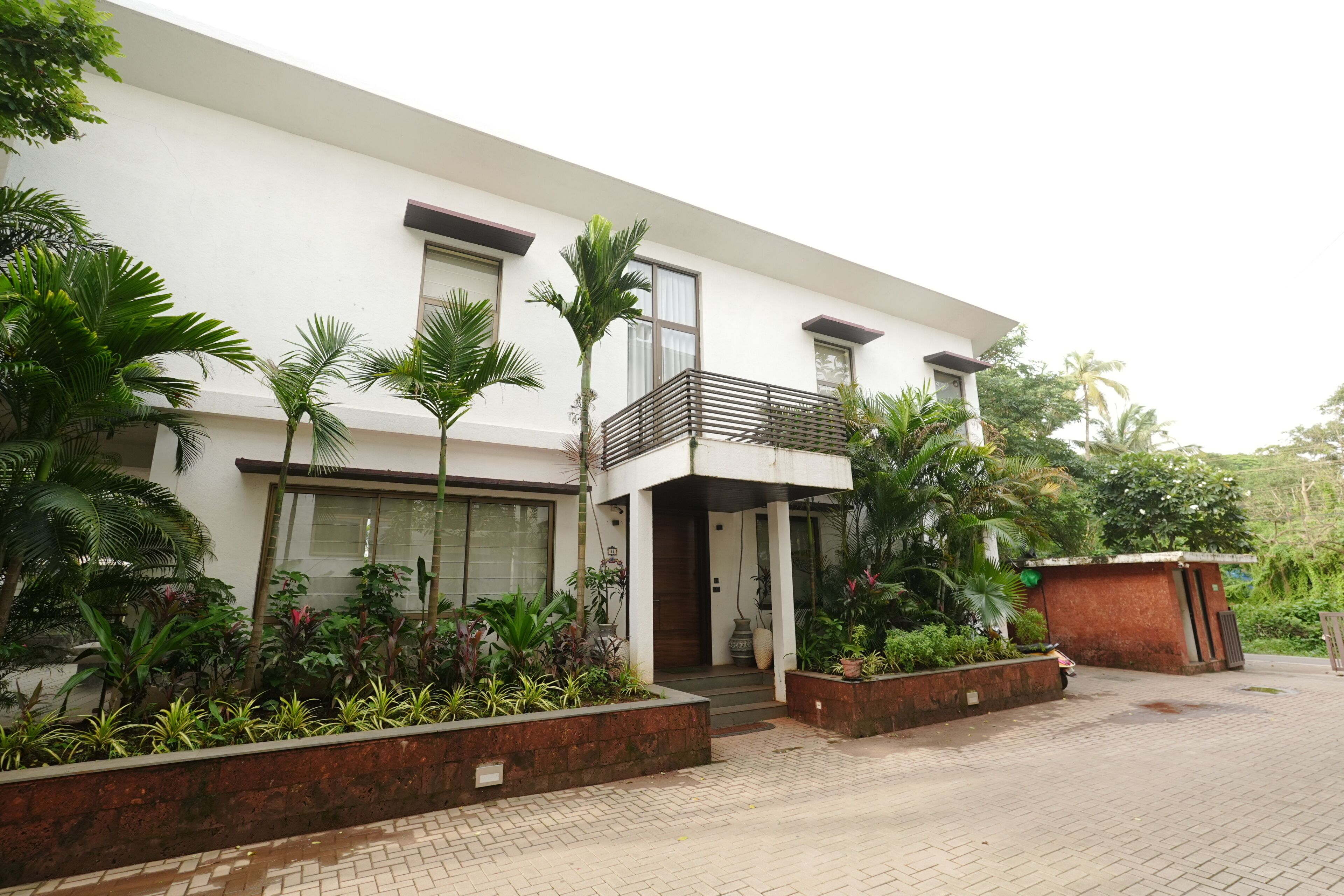 A modern two-story building with a balcony, surrounded by lush greenery and a paved driveway.