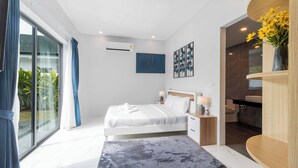 Ein modernes Schlafzimmer mit einem großen Bett, Nachttischen, einem kleinen Tisch, einem Badezimmer mit Waschbecken und Spiegel sowie Blick auf Grünflächen.