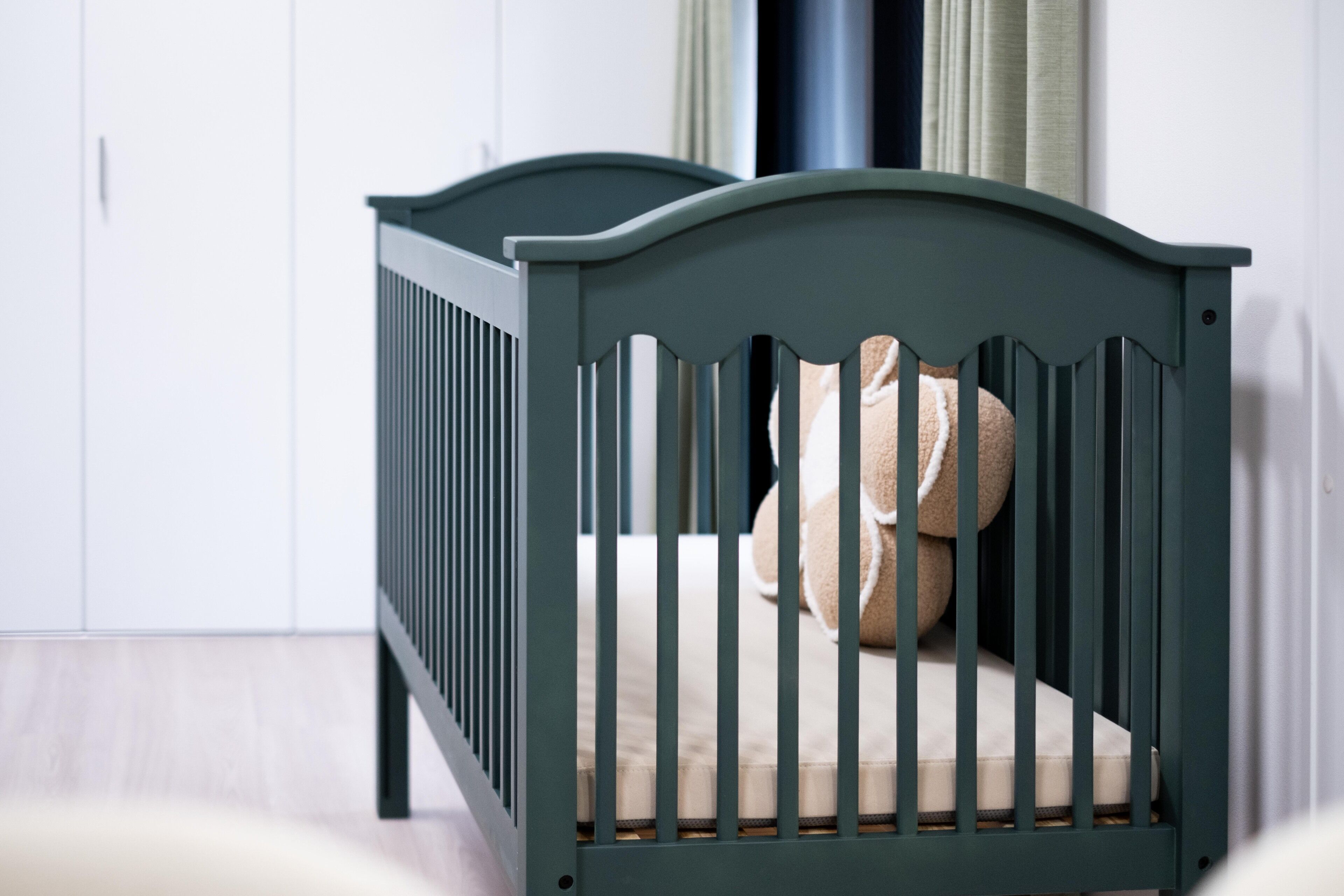 Un berceau vert foncé, doté d’un matelas moelleux et de deux oreillers, est placé contre un mur blanc orné d’une fenêtre et de rideaux.