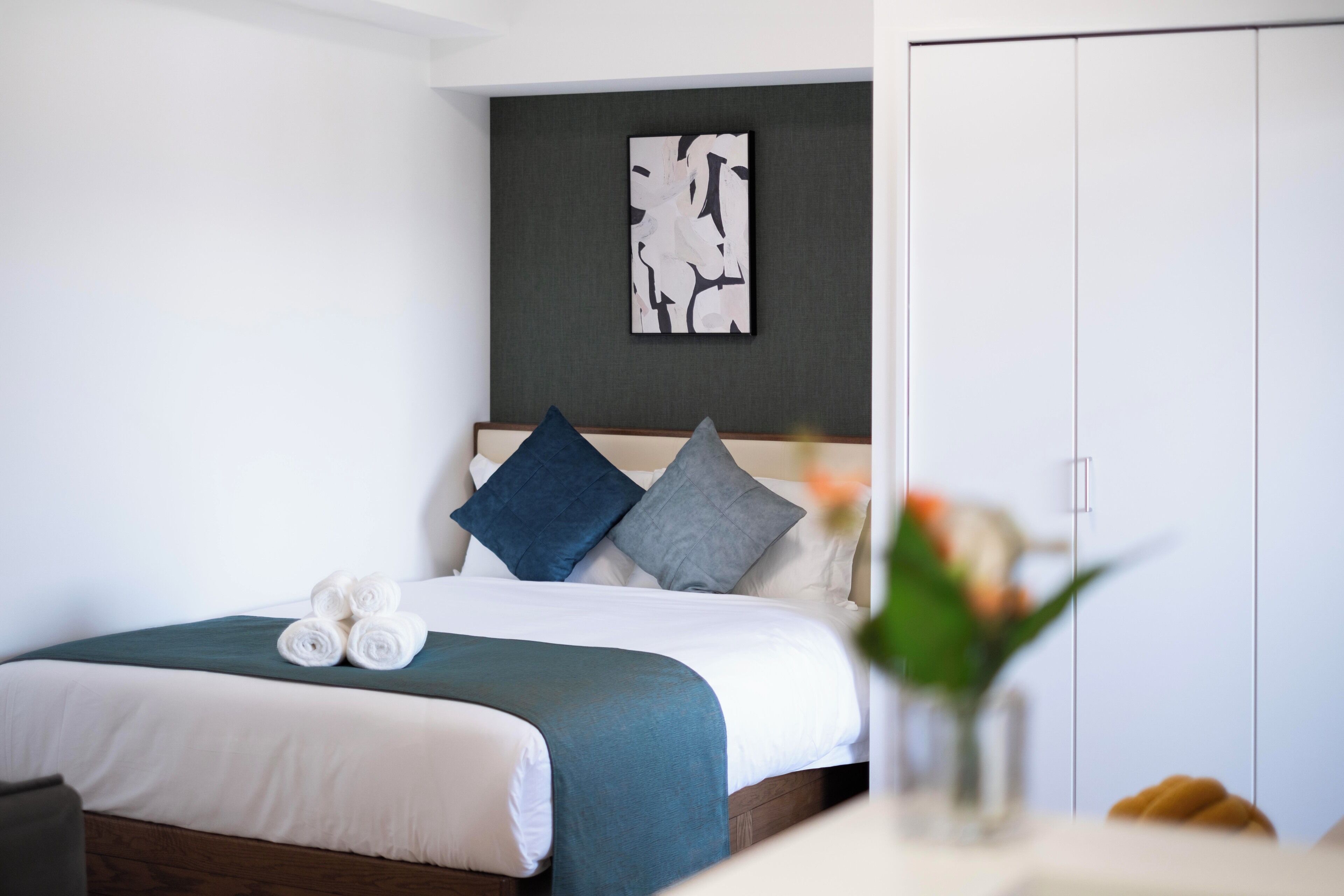 A neatly made bed with rolled towels, a dark headboard, and a wall art piece above it.