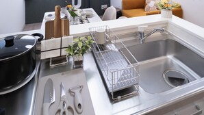 A modern kitchen with a stainless steel sink, a cutting board, and kitchen utensils. There is a dining area with a table and chairs, and a sofa in the background. - The Peak Bentencho (Osaka)