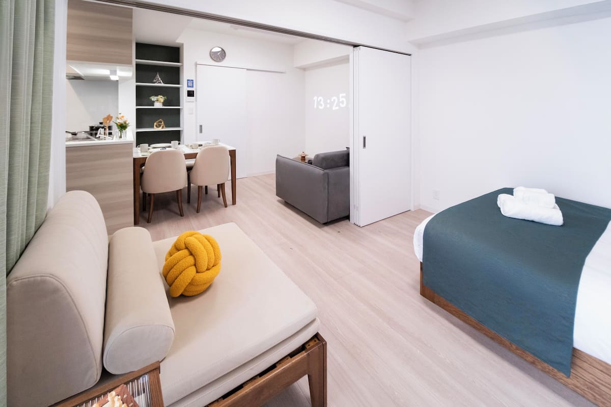 Quarto de hotel moderno com sofá, cama, escrivaninha com cadeiras, pequena cozinha e porta deslizante.