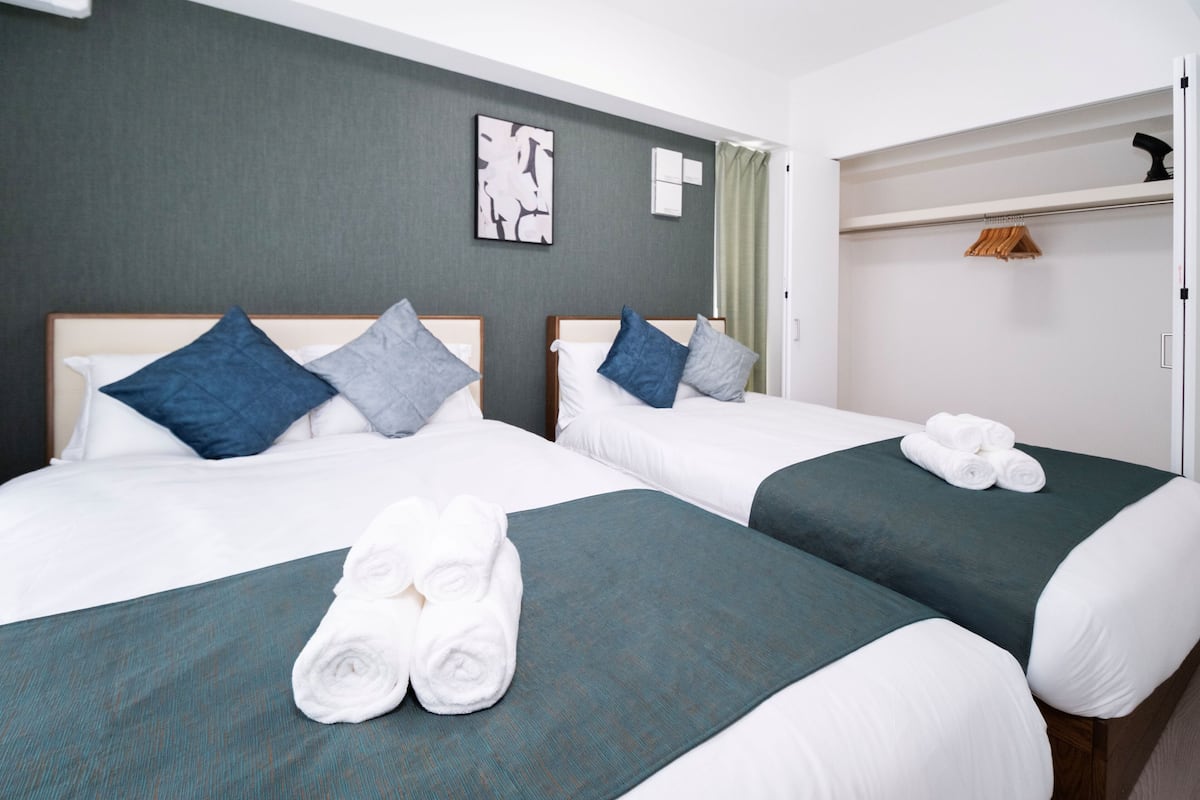 Quarto de hotel com duas camas, cada uma com colcha verde e roupa de cama branca, e um conjunto de toalhas brancas dobradas sobre a cama.