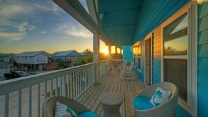 Una terraza de madera con sillas de mimbre y una pequeña mesa redonda, con vista a una playa y casas al fondo.