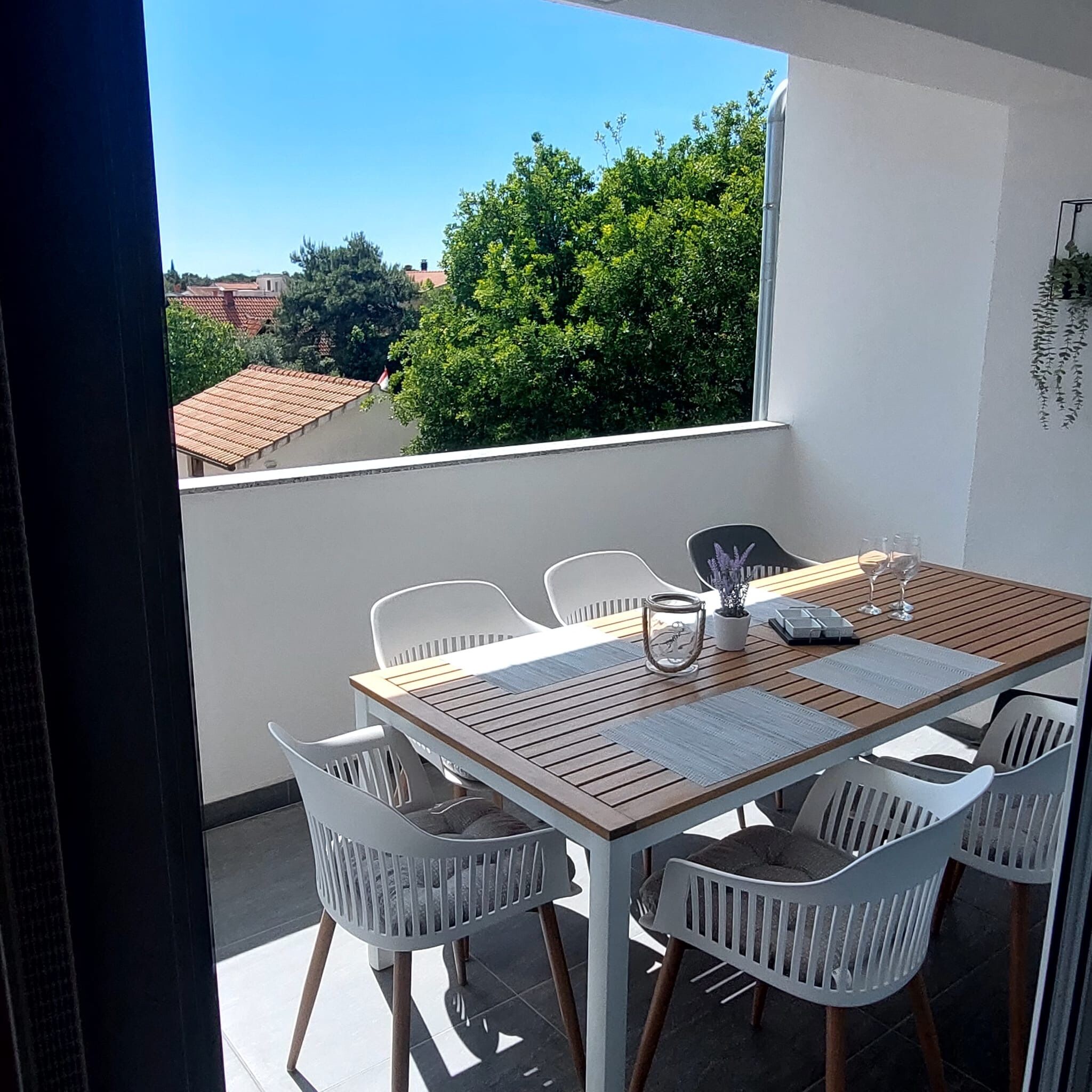 Een balkon met een houten tafel en witte stoelen, met uitzicht op een stadsgezicht met bomen en gebouwen.