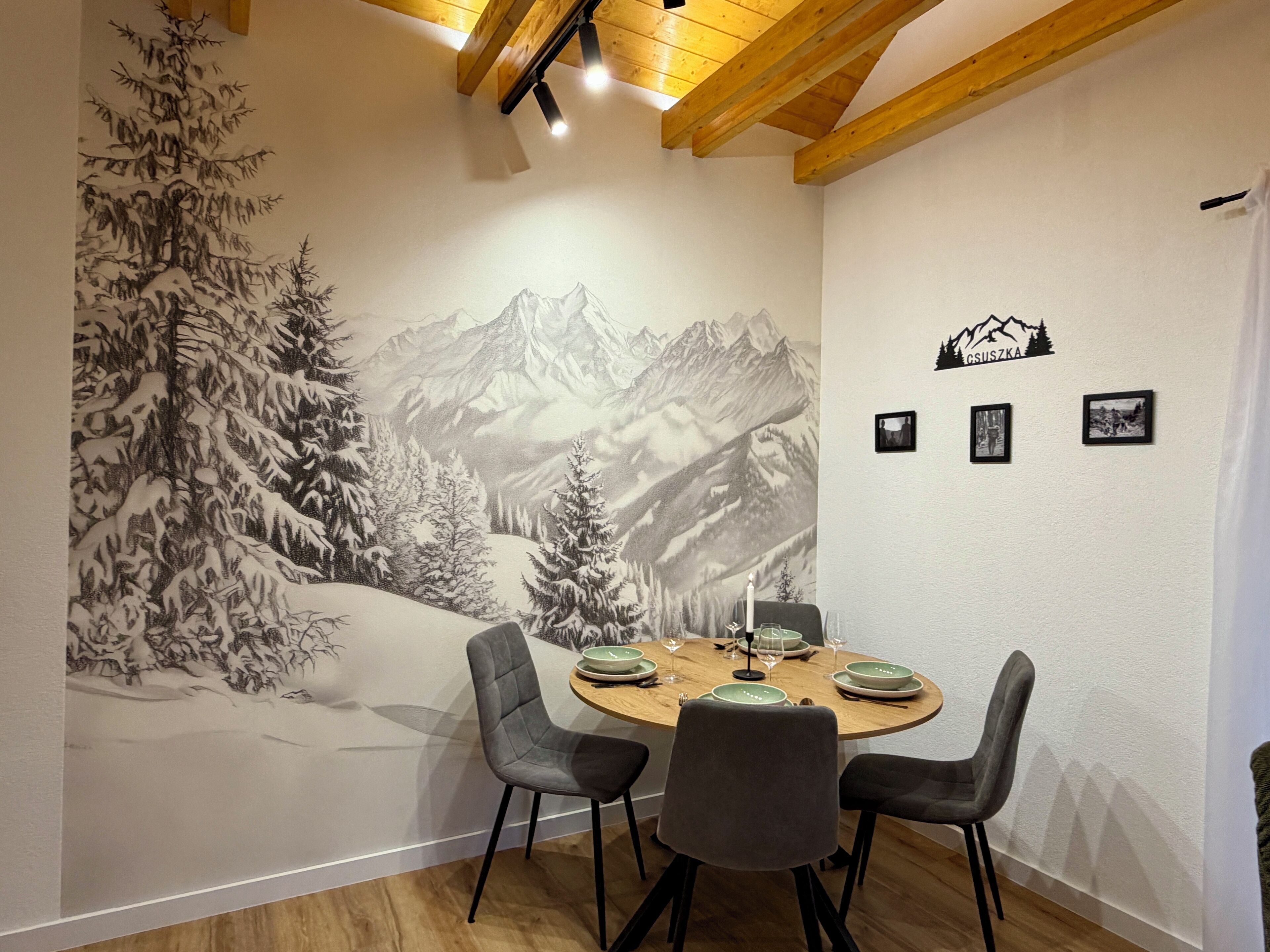 A dining area with a mural of a snowy mountain landscape, a round wooden table with four chairs, and three framed photographs on the wall.