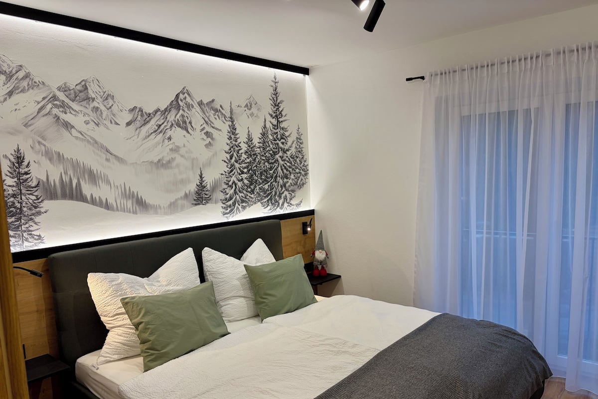 Ein Schlafzimmer mit Bett, Kissen, einem Nachttisch, einer Wandlampe und einem großen Gemälde einer Berglandschaft.