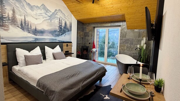 A bedroom with a large bed, a stone fireplace, a dining area with a table set for two, and a scenic mountain mural on the wall. - CSUSZKA VENDÉGHÁZ (Mátraszentimre)
