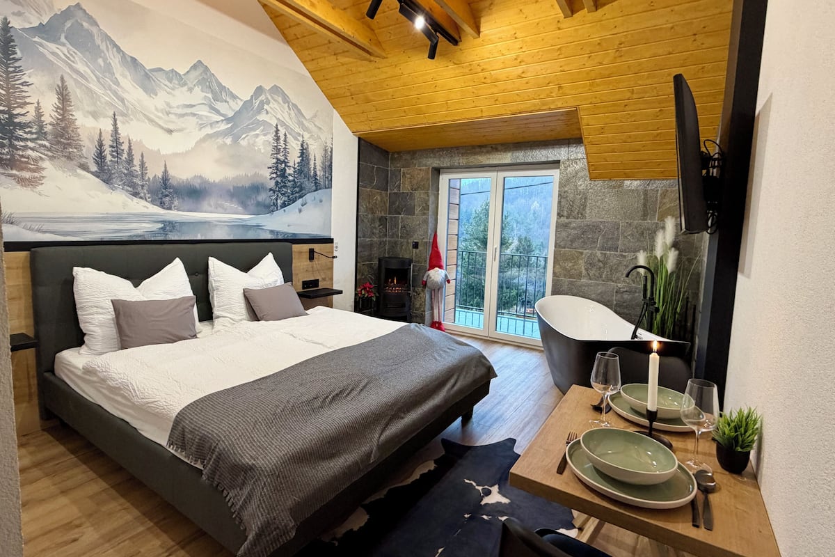 Ein Schlafzimmer mit einem großen Bett, einem Kamin aus Stein, einem Essbereich mit Tisch für zwei Personen und einem Wandgemälde mit einer Berglandschaft.
