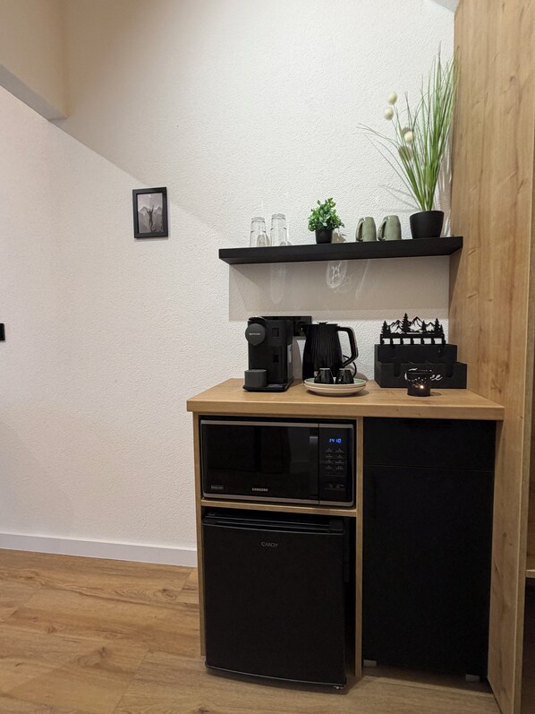 A compact kitchenette with a microwave, coffee maker, and a small refrigerator. There is a shelf above the counter holding a potted plant, a few mugs, and a decorative item. - CSUSZKA VENDÉGHÁZ (Mátraszentimre)
