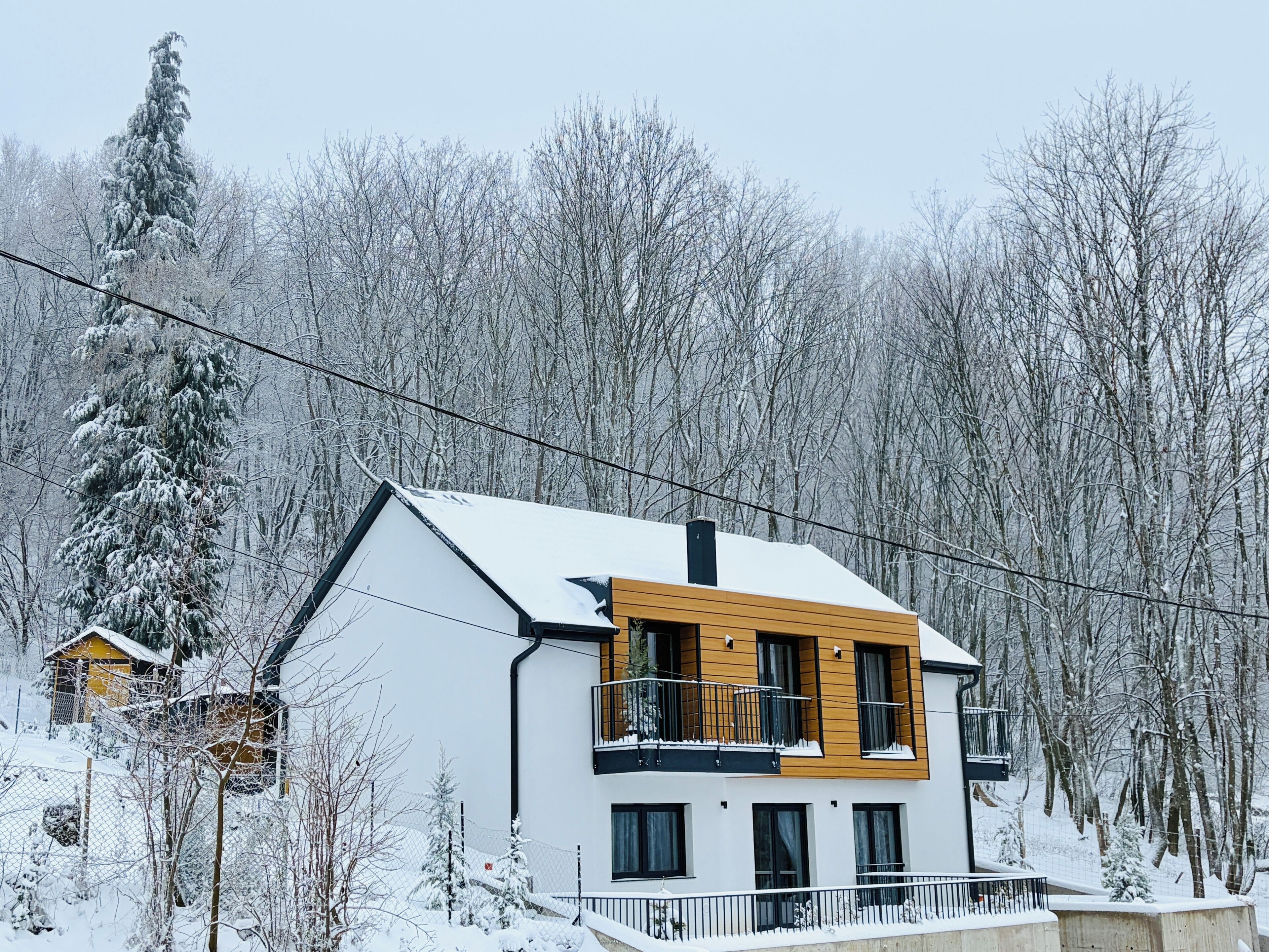 A modern two-story house with a balcony, surrounded by snow-covered trees.