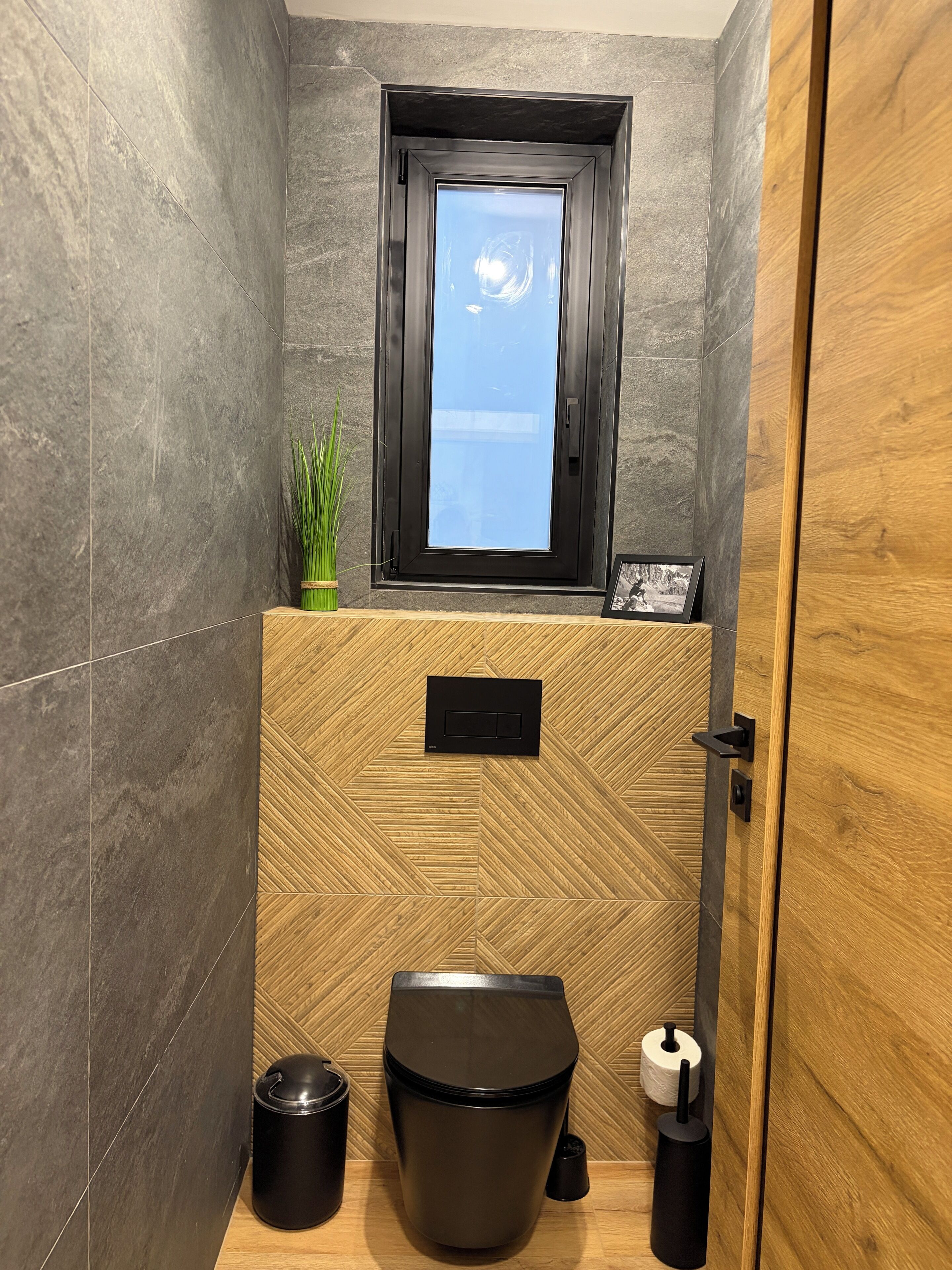 A modern bathroom with a window, a potted plant, a toilet, and a trash bin.