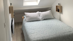 Une chambre avec un puits de lumière, un lit avec une literie à motifs, une petite table de chevet et une lampe fixée au mur.