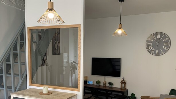 Un salon moderne avec une table à manger en bois, deux tabourets de bar, un grand miroir, un téléviseur et une horloge murale.