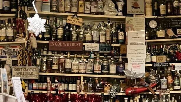 A bar area with shelves of liquor bottles, a sign reading "IN GOD WE TRUST," and a "SAVE WATER" sign. - Accomodation in House (Kinvara)
