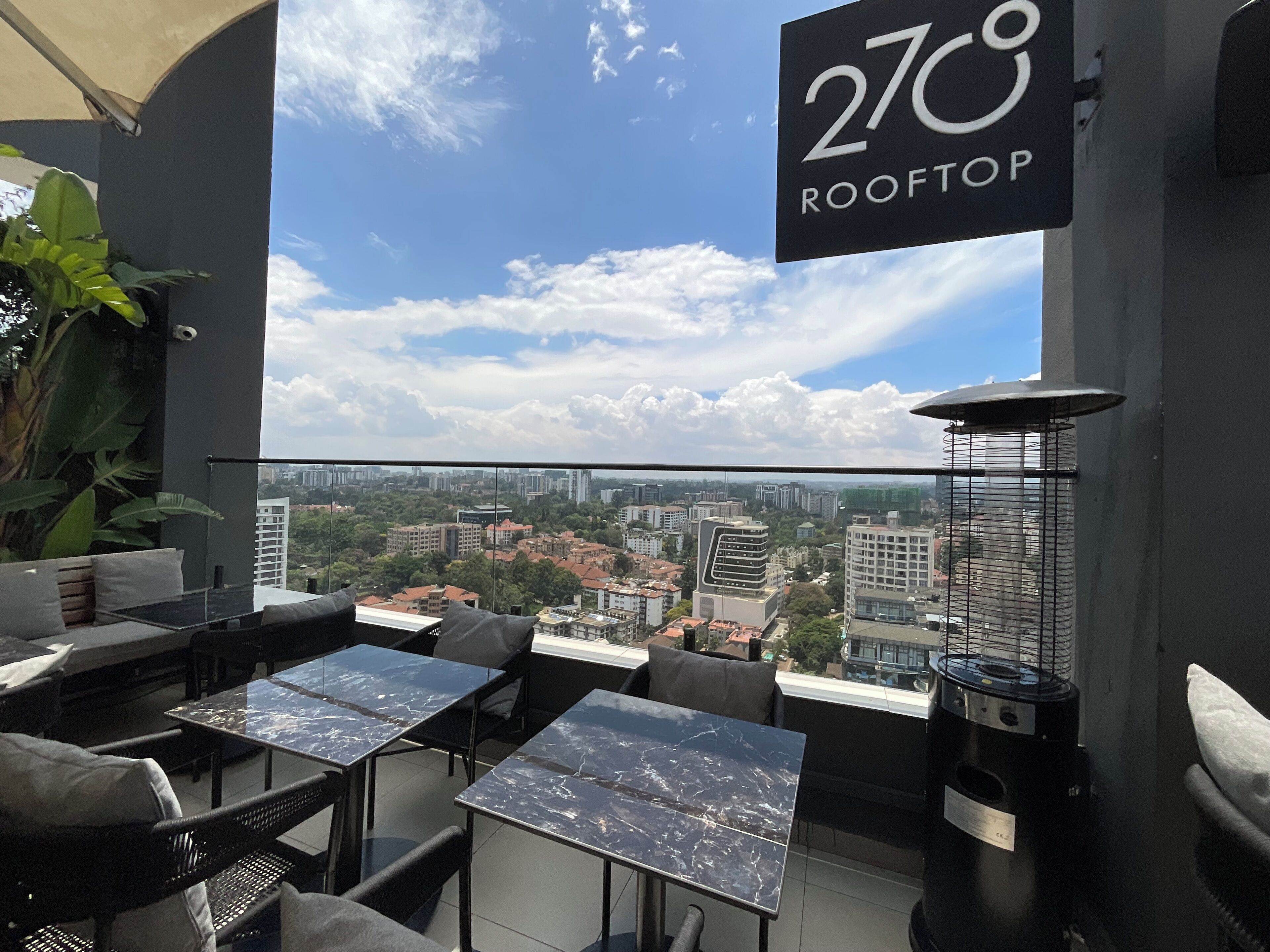 Un bar en la azotea con mesas al aire libre, vistas despejadas del paisaje urbano y un cartel que indica una temperatura de 27°C.