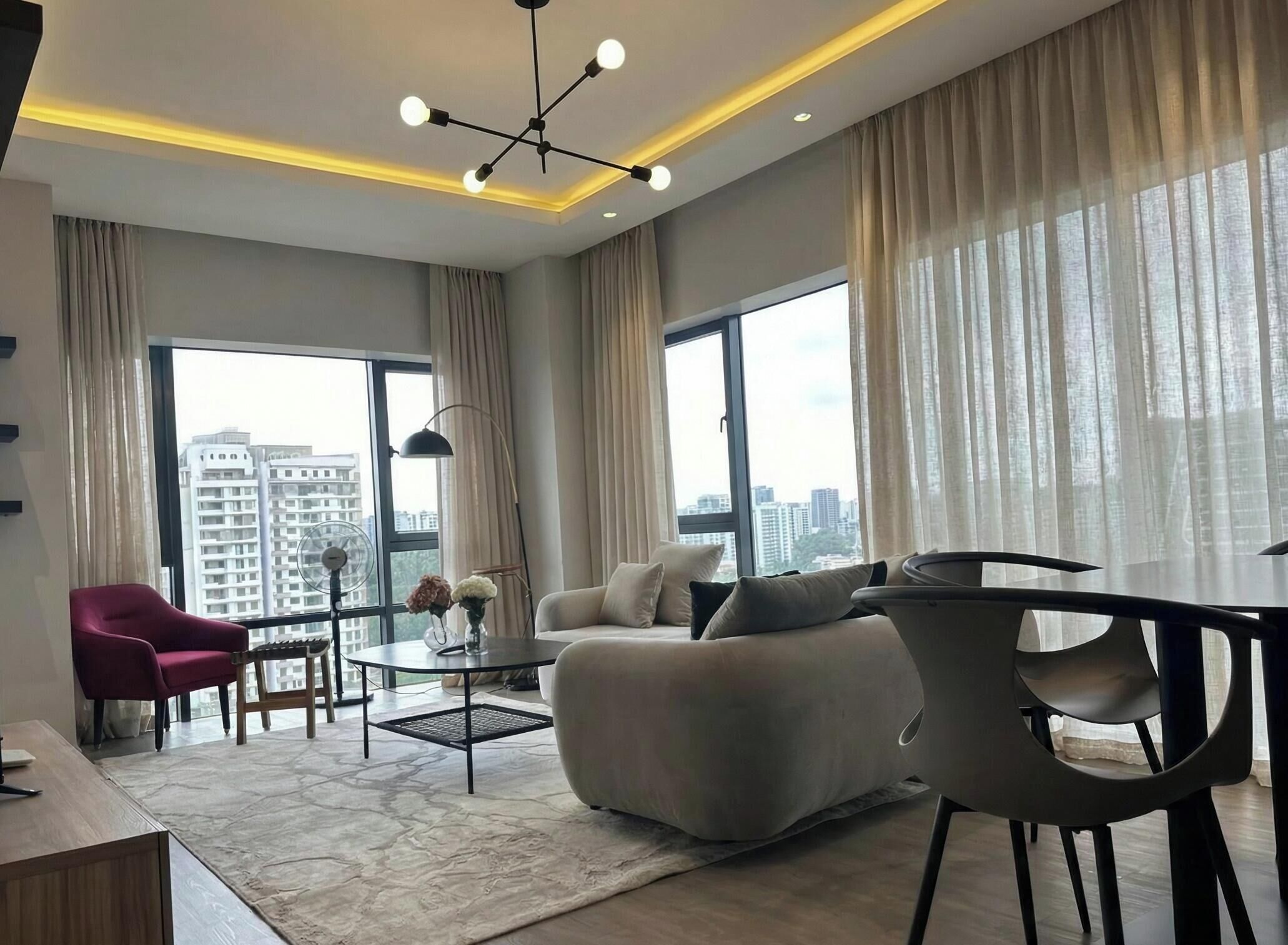 A modern living room with a sofa, a red chair, a dining table, and a large window with a city view.