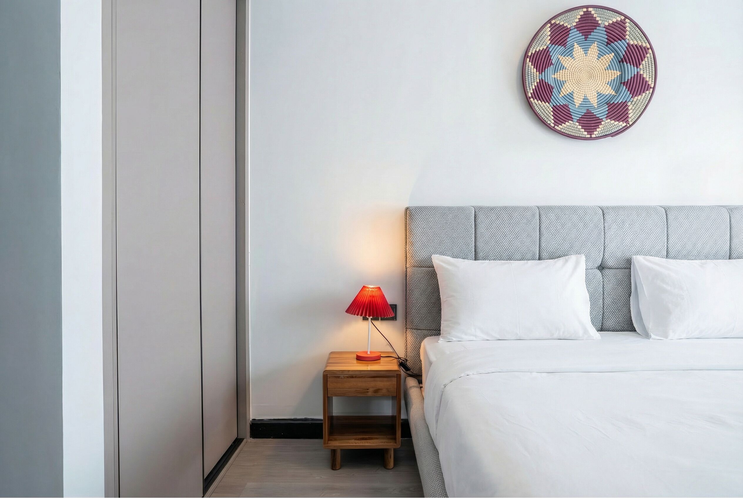 A neatly made bed with white bedding, a grey upholstered headboard, and a small wooden nightstand with a red lamp.