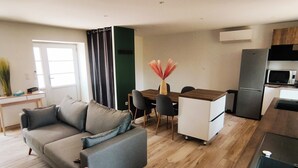 Una sala de estar moderna con un sofá gris, una mesa de comedor con sillas, un área de cocina con refrigerador y microondas, y piso de madera.